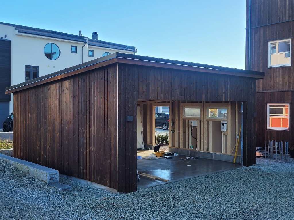 Moderne mørk trekledd carport under arbeid med synlig kabling og veggmontert elbillader.
